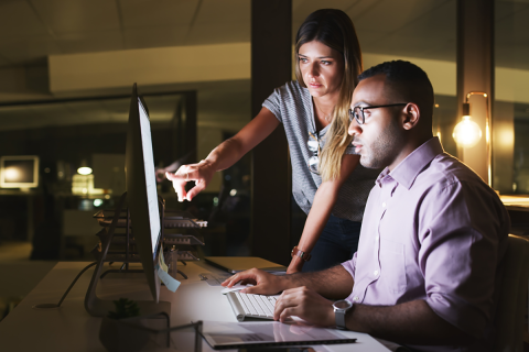 Two corporate employees, a man and woman, looking at a computer monitor together, with the woman pointing at the screen.