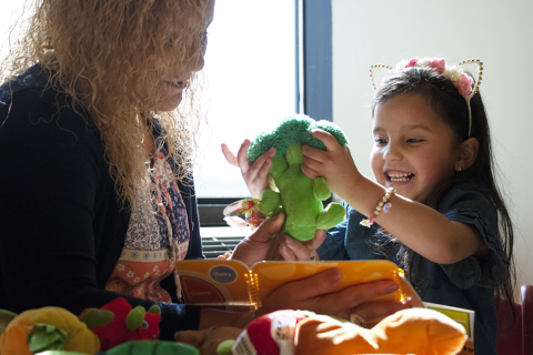 A woman reading a child a book, as the child is playing with a broccoli plush toy.